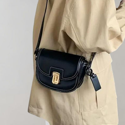 Sac à bandoulière noir en cuir avec fermoir doré, porté par une personne en manteau beige. Accessoire mode élégant et tendance.