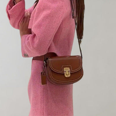 Femme en tailleur rose portant un sac à main marron en cuir avec bandoulière, sur fond neutre. Mode élégante, accessoires tendance.