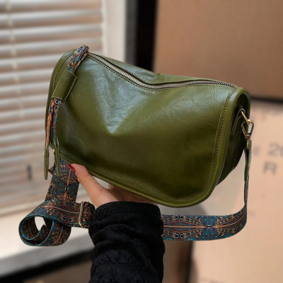 Sac à bandoulière vert olive en cuir avec fermeture éclair, porté par une main. Sangle décorative colorée. Accessoire de mode tendance.