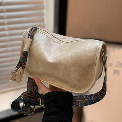 Sac à bandoulière beige en cuir synthétique avec sangle décorative colorée, tenu par une main. Accessoire mode tendance pour femme.