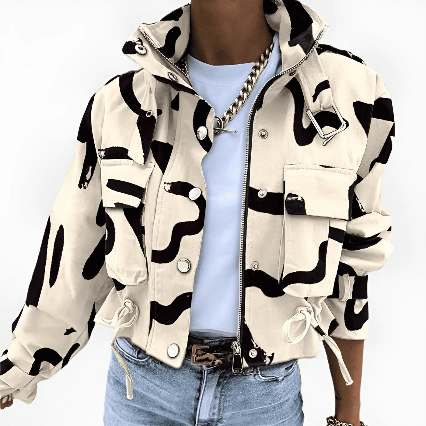 Veste camouflage beige et noire, style militaire, avec poches et cordons, portée par une personne en jean et t-shirt blanc, chaîne dorée visible.