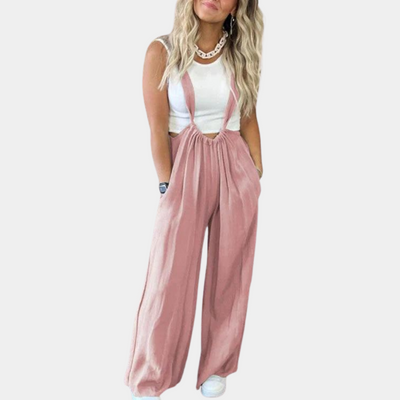 Femme portant une salopette rose ample avec un haut blanc sans manches, cheveux blonds ondulés, posant sur fond blanc. Mode décontractée chic.