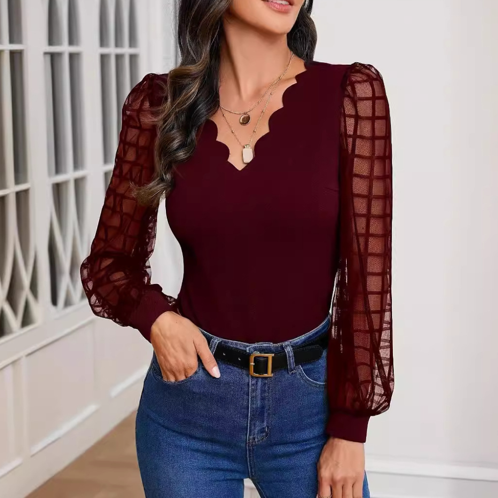 Femme portant un haut bordeaux à manches longues transparentes et col festonné, avec un jean bleu et ceinture noire, dans un intérieur moderne.