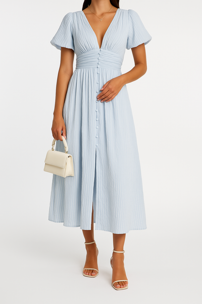 Robe longue bleue à manches courtes, décolleté en V, boutons devant. Femme tenant un sac à main blanc, portant des sandales à talons. Mode élégante.
