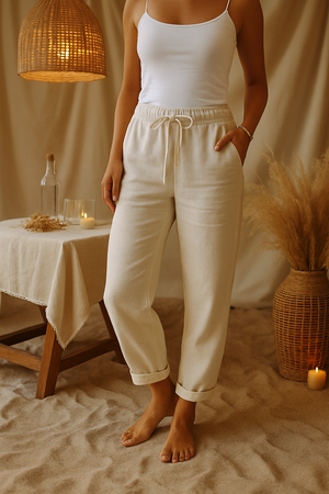 Femme en débardeur blanc et pantalon beige, debout sur du sable intérieur, décor bohème avec lampe suspendue, table en bois et vase en osier.