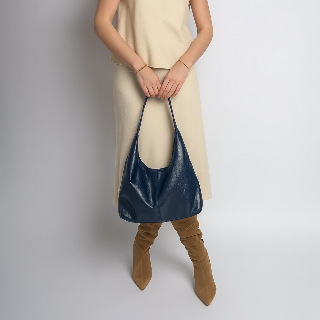 Femme en robe beige tenant un sac à main bleu en cuir, portant des bottes marron. Mode élégante, accessoires tendance, style automne-hiver.