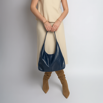 Femme en robe beige tenant un sac à main bleu en cuir, portant des bottes marron. Mode élégante, accessoires tendance, style automne-hiver.