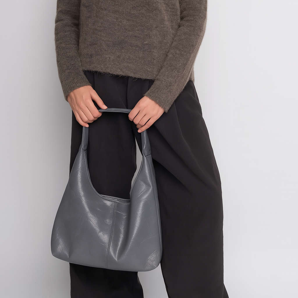 Femme tenant un sac à main en cuir gris, vêtue d'un pull marron et d'un pantalon noir. Mode élégante, accessoires tendance, style automne-hiver.