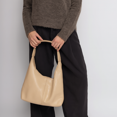 Femme tenant un sac à main beige en cuir, vêtue d'un pull marron et d'un pantalon noir. Mode élégante, accessoires tendance, style automne-hiver.