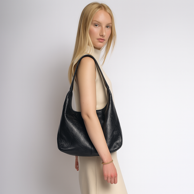Femme portant un sac à main en cuir noir élégant, robe beige, fond blanc. Mode féminine, accessoire tendance, style chic et moderne.