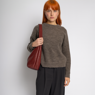 Jeune femme rousse portant un pull marron et un sac à main en cuir rouge, posant contre un fond blanc. Mode automne, style décontracté élégant.