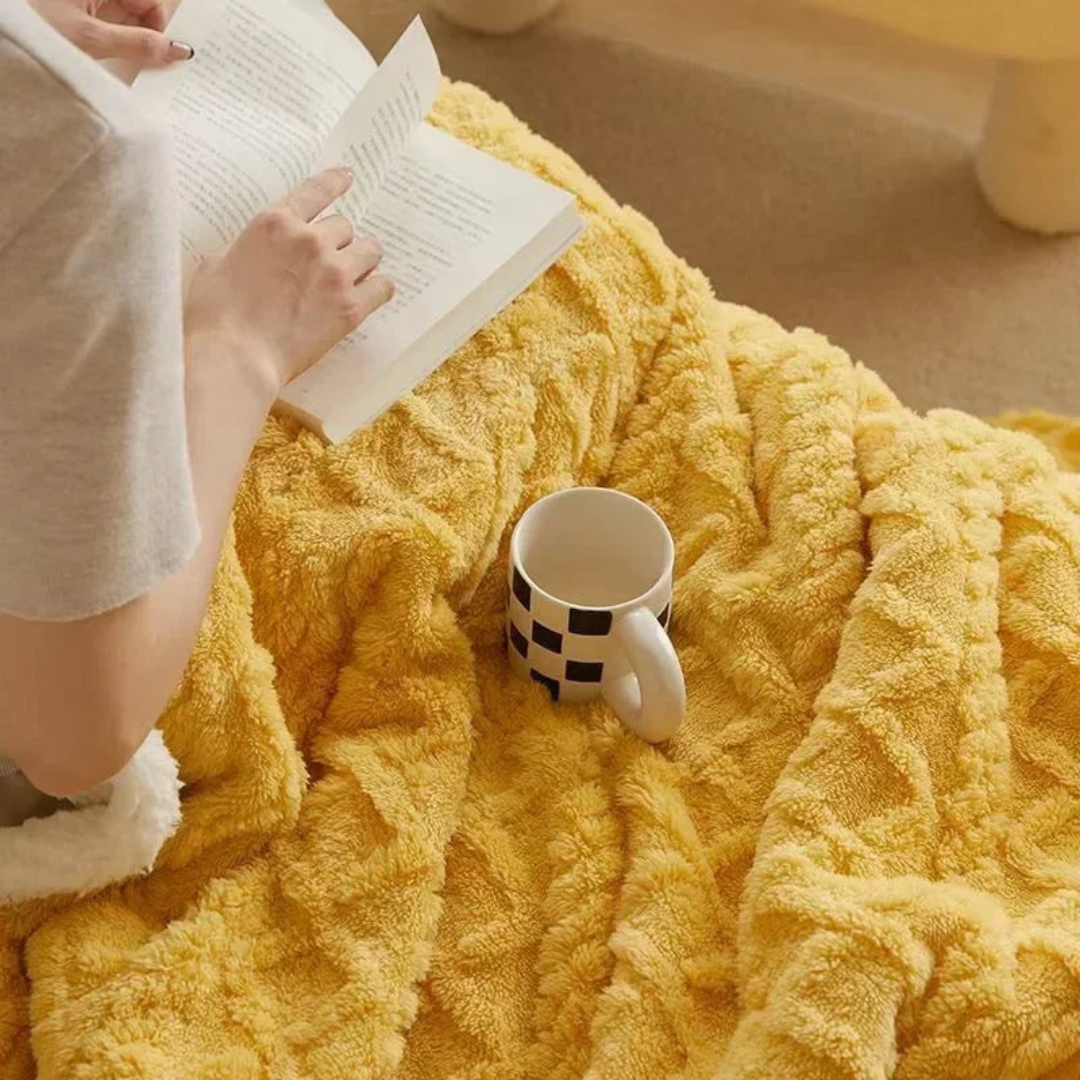Personne lisant un livre sur une couverture jaune moelleuse avec une tasse à motifs noirs et blancs. Moment de détente, lecture, confort à la maison.