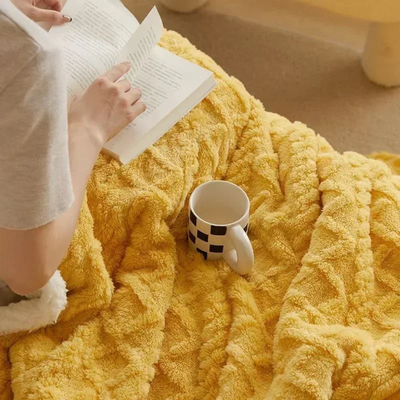 Personne lisant un livre sur une couverture jaune moelleuse avec une tasse à motifs noirs et blancs. Moment de détente, lecture, confort à la maison.