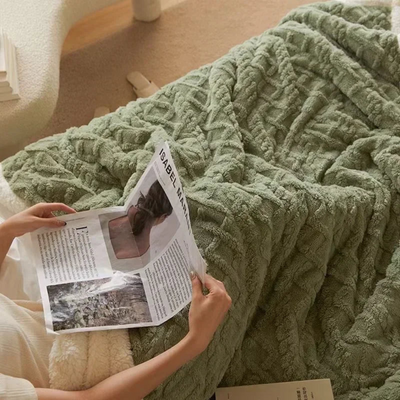 Personne lisant un magazine sur un canapé, recouverte d'une couverture en fausse fourrure verte. Ambiance cosy, détente à la maison.