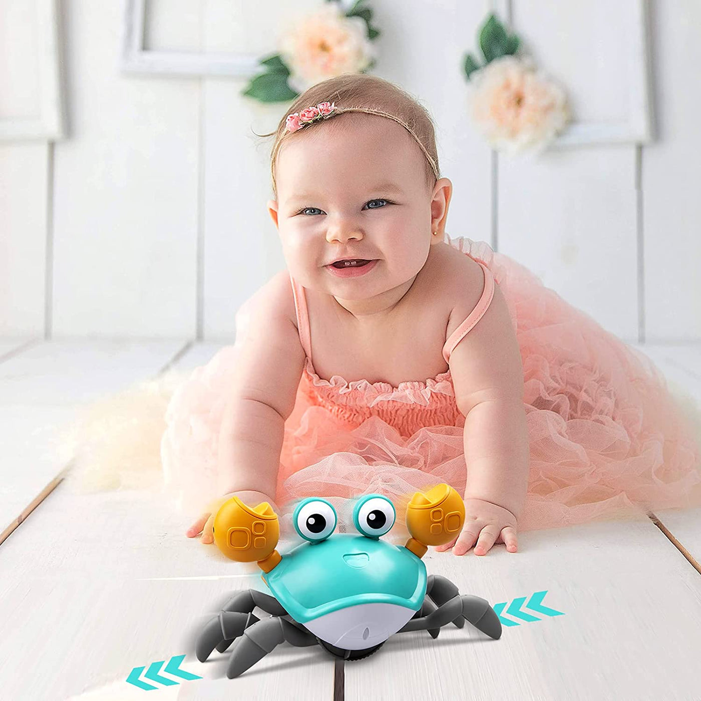 Bébé souriant en robe rose avec jouet crabe interactif bleu sur sol blanc, décor floral en arrière-plan. Jouet éducatif, cadeau bébé.