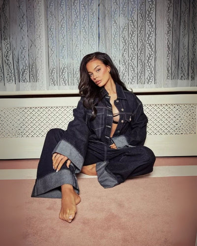 Jeune femme posant en tenue en jean oversize, assise sur un tapis rose devant un rideau blanc. Mode décontractée, style denim tendance.