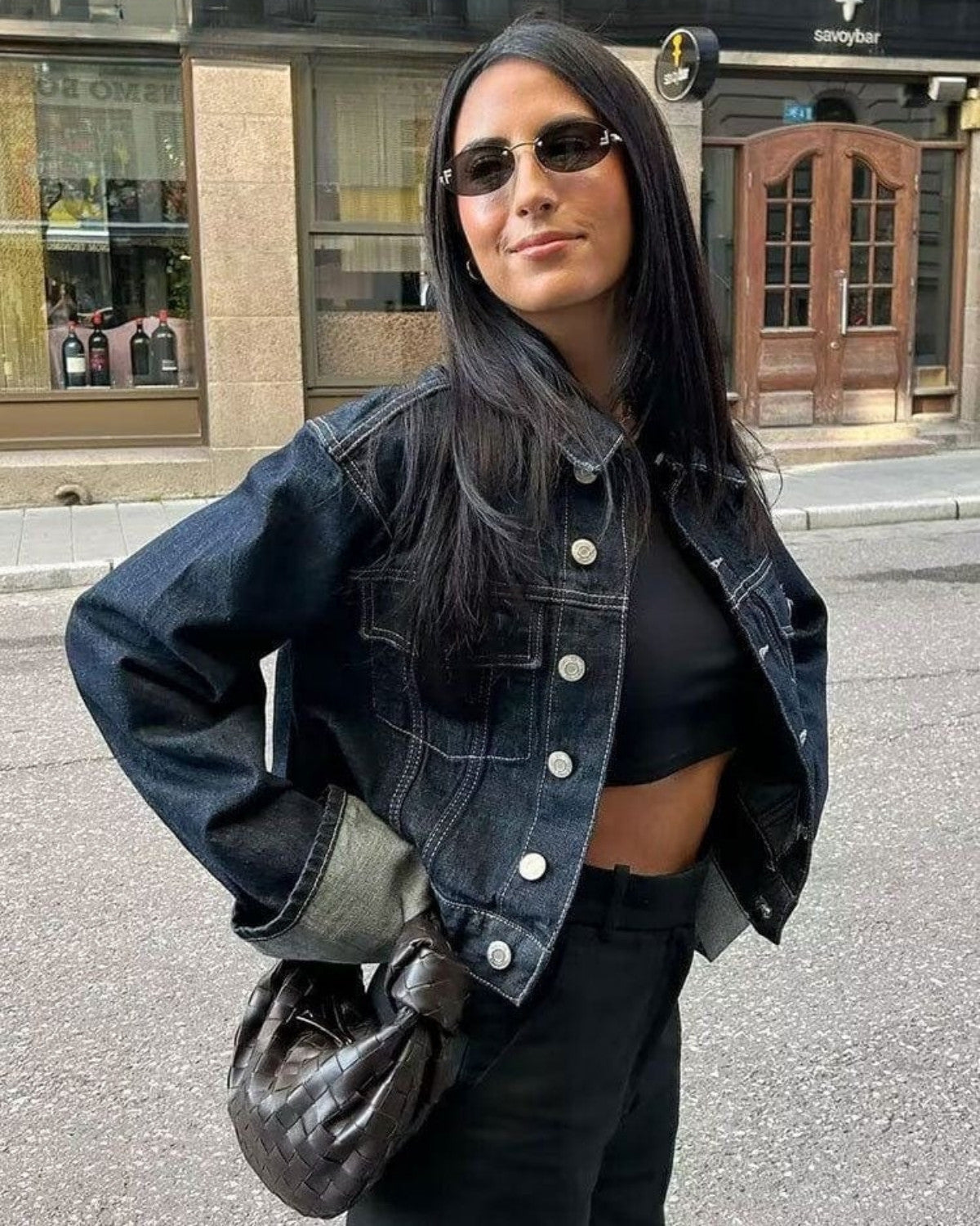 Femme portant une veste en jean, lunettes de soleil noires, sac tressé marron, posant dans une rue urbaine. Mode décontractée, style moderne.