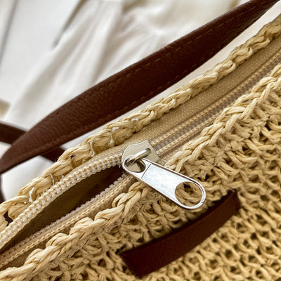 Sac en paille tressée avec fermeture éclair argentée et anses en cuir marron, posé sur un fond blanc. Accessoire mode estival élégant.
