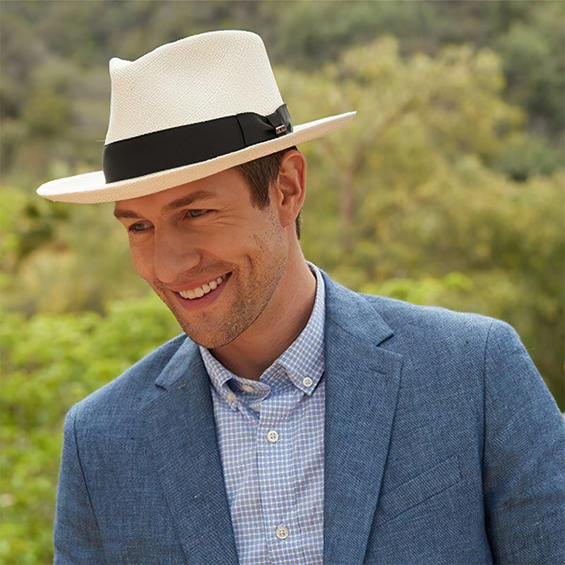 Homme souriant portant un chapeau Panama blanc avec ruban noir, costume bleu et chemise à carreaux, en extérieur verdoyant. Mode élégante masculine.