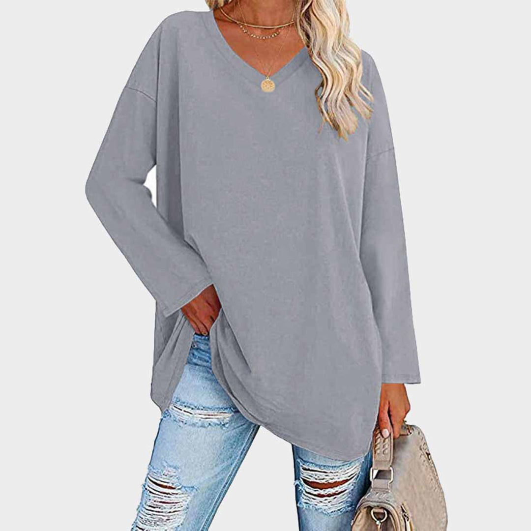 Femme portant un pull gris ample à col en V, associé à un jean déchiré et un sac beige. Mode décontractée, tendance automne-hiver.