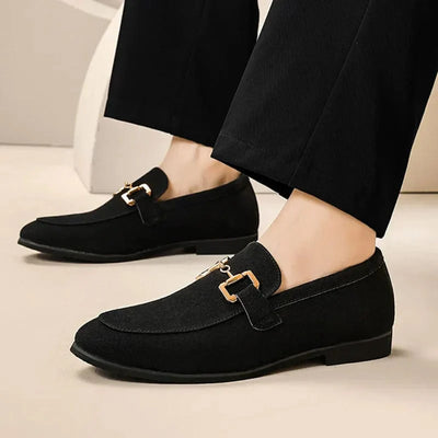 Chaussures mocassins noirs élégants pour homme avec boucle dorée, portés avec un pantalon noir. Style moderne et chic pour tenues formelles.