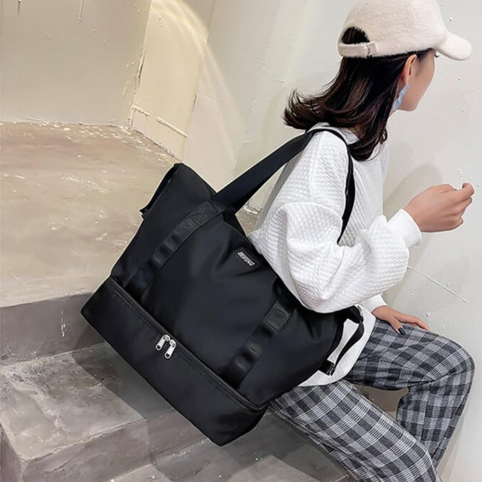 Femme assise sur des marches, portant un sac de sport noir spacieux, tenue décontractée avec casquette blanche et pantalon à carreaux.