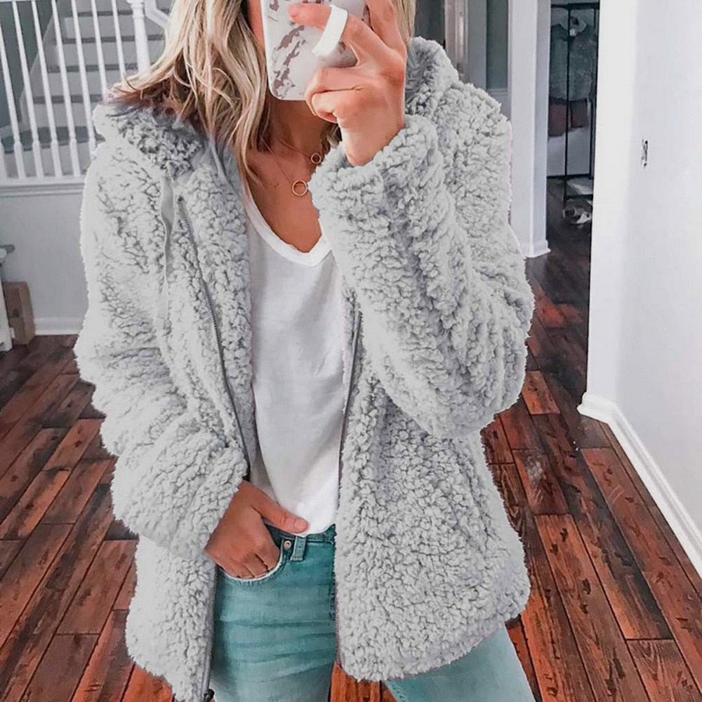 Femme portant un manteau en fausse fourrure gris, t-shirt blanc et jean, tenant un smartphone, sur un sol en bois. Mode hiver décontractée.