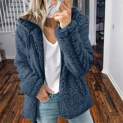 Femme portant une veste en fausse fourrure bleue, t-shirt blanc et jean, debout sur un sol en bois. Mode hiver, style décontracté, intérieur maison.