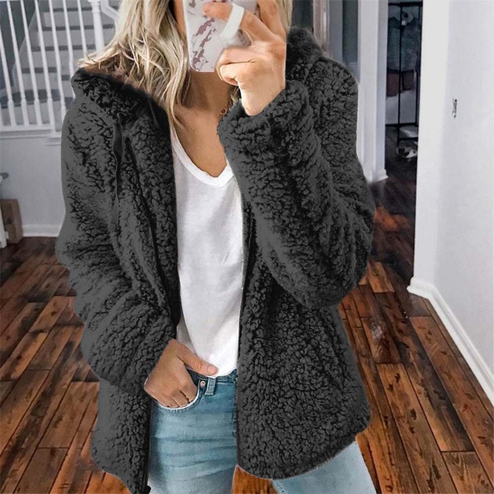 Femme portant une veste en fausse fourrure noire, t-shirt blanc et jean, tenant un smartphone, debout sur un sol en bois dans un intérieur moderne.