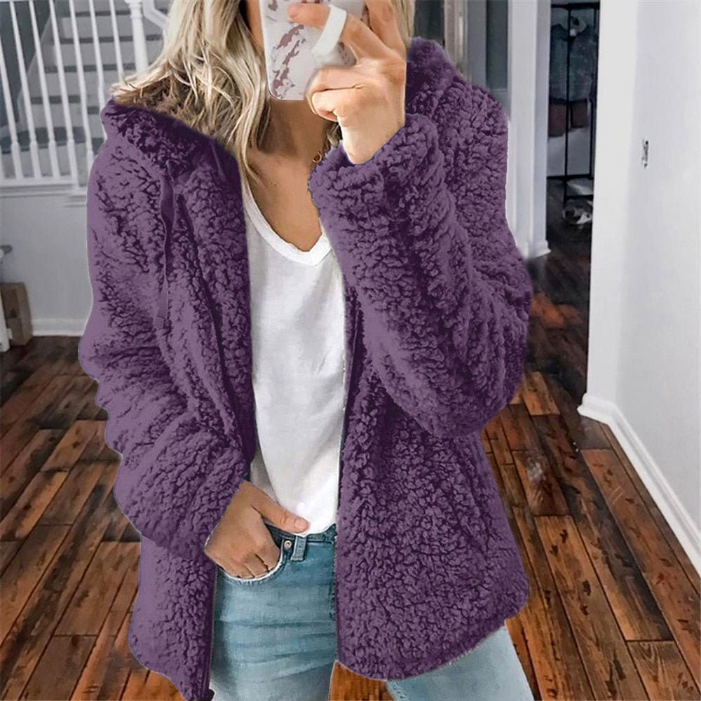 Femme portant une veste en fausse fourrure violette, debout sur un sol en bois, tenant un smartphone. Mode hiver, style décontracté, tendance.
