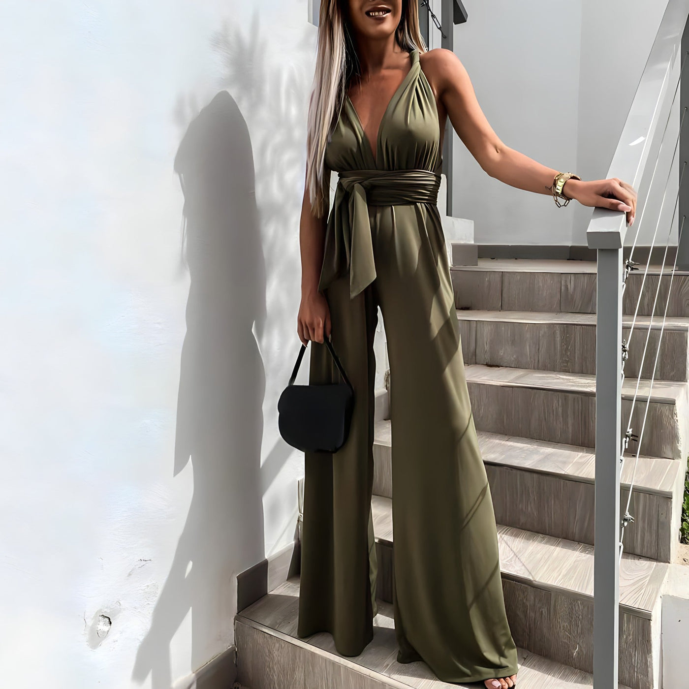 Femme en combinaison verte élégante sur escalier moderne, tenant un sac noir. Mode féminine chic, tenue de soirée tendance, style urbain.