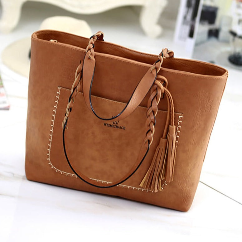 Sac à main en cuir marron avec anses tressées et pompons, posé sur une table blanche. Accessoire mode élégant et tendance pour femme.