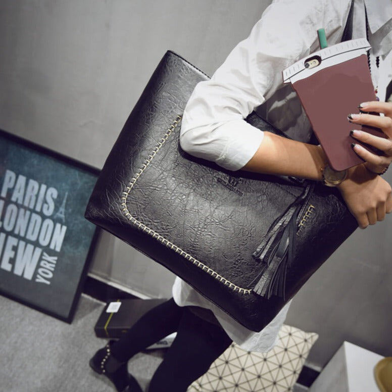 Sac à main noir élégant avec coutures apparentes, porté par une personne en chemise blanche, tenant un gobelet. Décor moderne en arrière-plan.