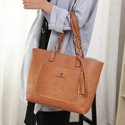 Sac à main en cuir marron avec anses tressées, pompons décoratifs, porté par une personne en chemise grise. Accessoire mode élégant et tendance.