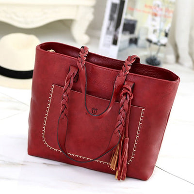 Sac à main en cuir rouge avec tresses, poche avant et pompons, posé sur une surface blanche. Accessoire mode élégant et tendance.