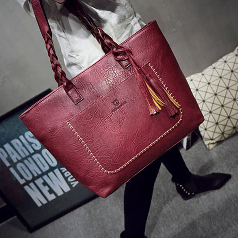 Sac à main en cuir rouge bordeaux avec anses tressées, détails de couture, et pompons, posé sur un fond moderne. Accessoire mode élégant.