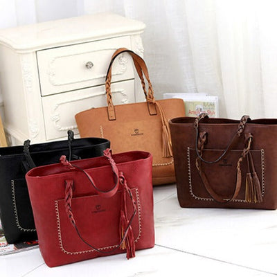 Quatre sacs à main en cuir de différentes couleurs (noir, rouge, marron) posés devant une commode blanche, avec des détails tressés et pompons.