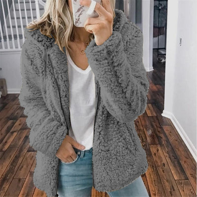 Femme portant une veste en fausse fourrure grise, t-shirt blanc et jean, tenant un smartphone. Mode automne-hiver, style décontracté.