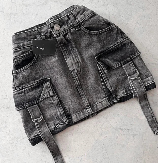 Jupe en jean noir délavé avec poches cargo et ceinture, posée sur fond gris. Mode urbaine, tendance streetwear, style décontracté.