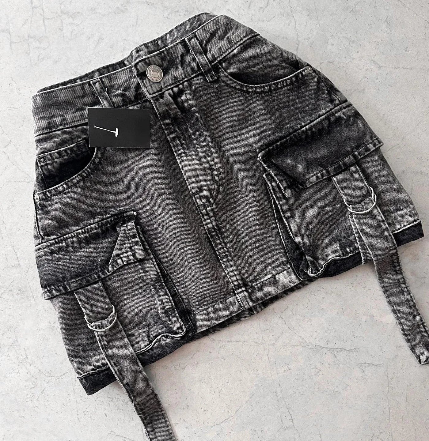 Jupe en jean noir délavé avec poches cargo et ceinture, posée sur fond gris. Mode urbaine, tendance streetwear, vêtement femme moderne.
