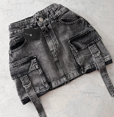 Jupe en jean noir délavé avec poches cargo et ceinture, posée sur fond gris. Mode urbaine, tendance streetwear, vêtement femme moderne.