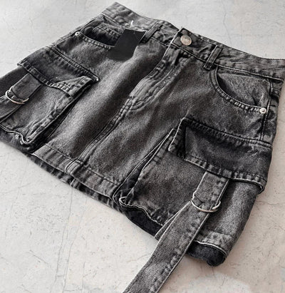 Jupe en jean noir avec poches cargo et ceinture, posée sur une surface en béton. Mode urbaine, tendance streetwear, style décontracté.