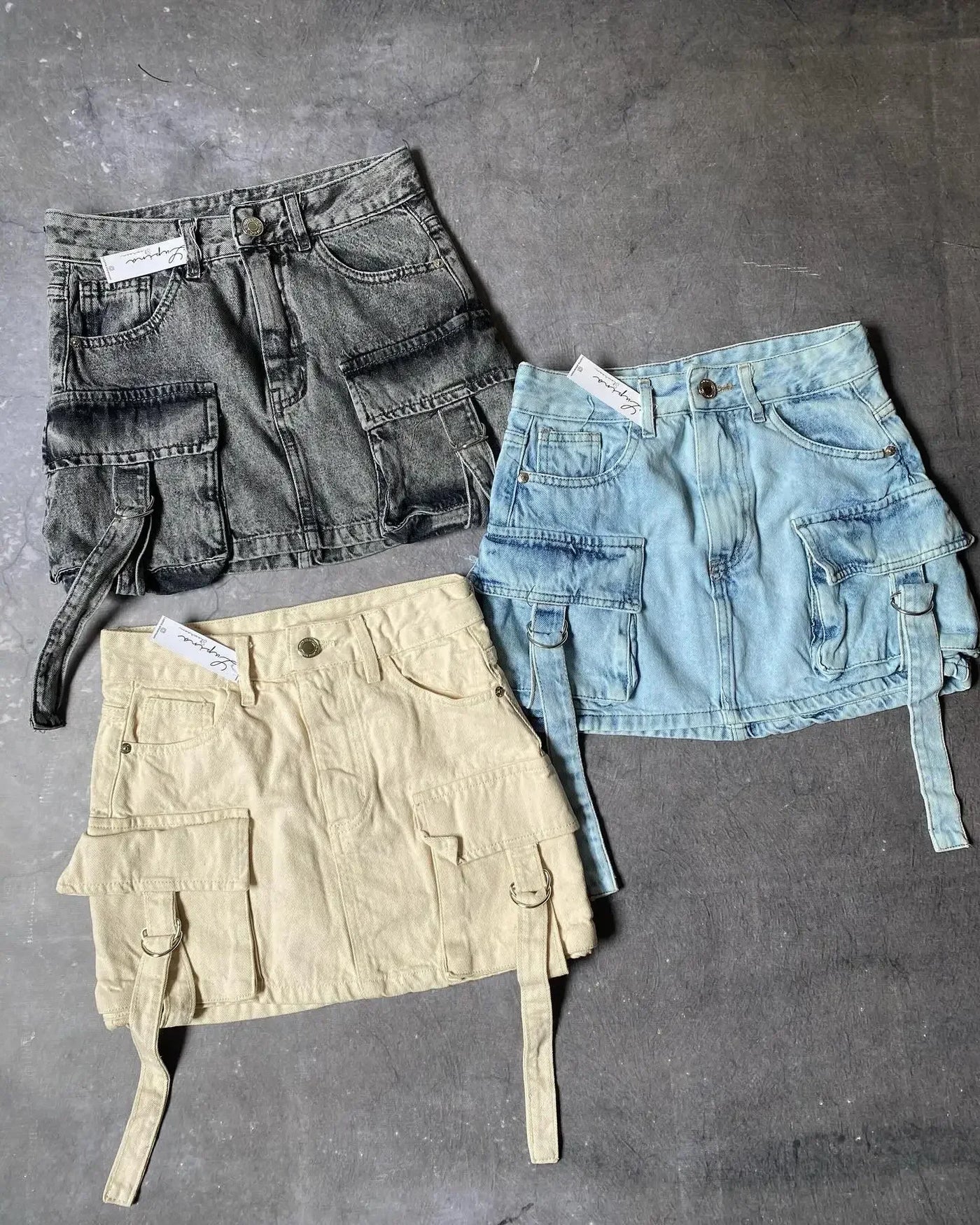 Trois jupes cargo en denim sur fond gris: une noire, une bleue et une beige, avec poches multiples et ceintures, style tendance et urbain.