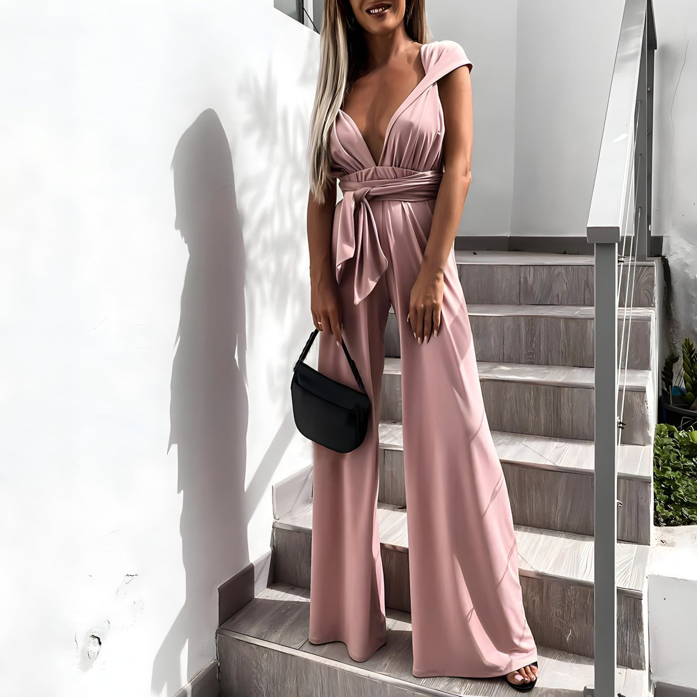Femme en combinaison rose élégante, tenant un sac à main noir, posant sur des escaliers modernes. Mode féminine, tenue chic, style urbain.