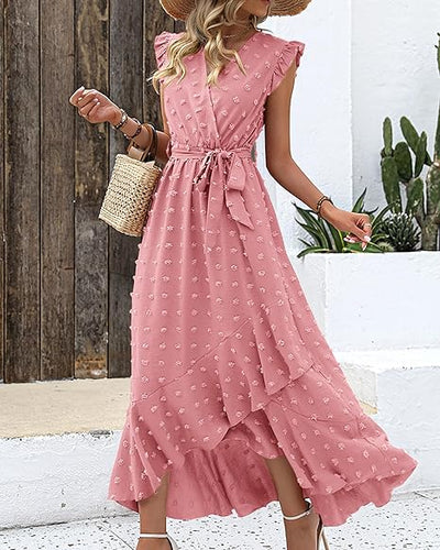 Robe longue rose à volants avec motifs floraux, portée par une femme tenant un sac en osier, devant un mur en bois et des plantes succulentes.
