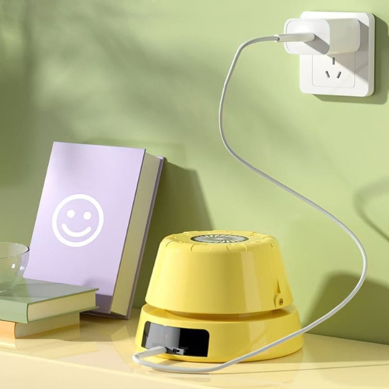 Chargeur mural blanc branché, câble connecté à un ventilateur jaune sur une table, livre violet avec smiley, mur vert en arrière-plan.
