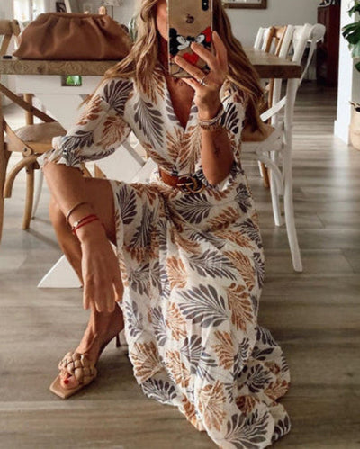 Robe longue à motifs floraux beige et gris, ceinturée, portée par une femme prenant un selfie. Chaussures tressées et sac à main marron en arrière-plan.