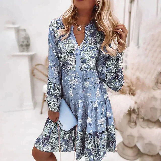 Robe fleurie bleue à manches longues, femme blonde tenant une pochette bleue, style bohème chic, intérieur lumineux, mode tendance.