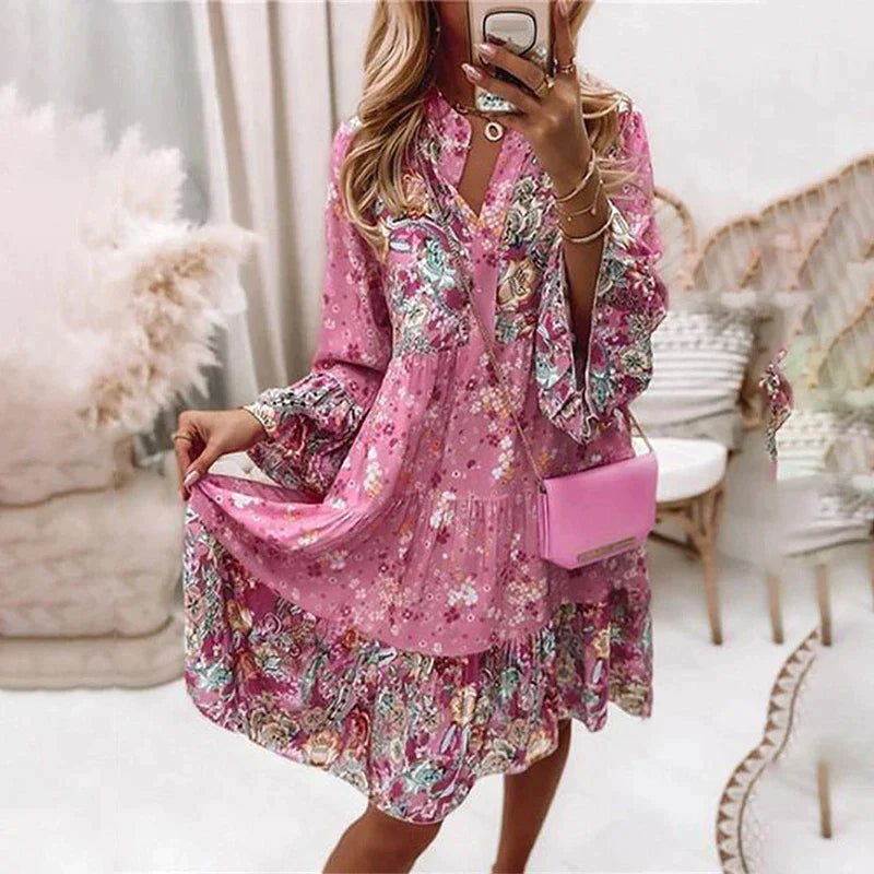 Robe rose à motifs floraux portée par une femme, avec un sac à main rose. Style bohème chic, manches longues, ambiance intérieure élégante.