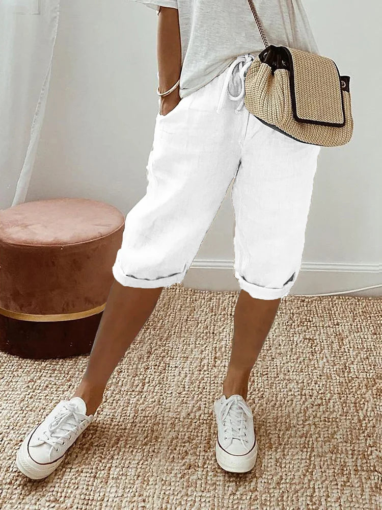 Femme portant un short blanc en lin, t-shirt gris, baskets blanches et sac à main en paille, posant sur un tapis beige. Mode décontractée estivale.